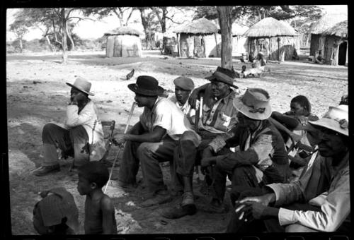 Group of men sitting and watching a dance; huts in the background