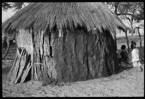 Woman and another person sitting beside a hut, seen from a distance
