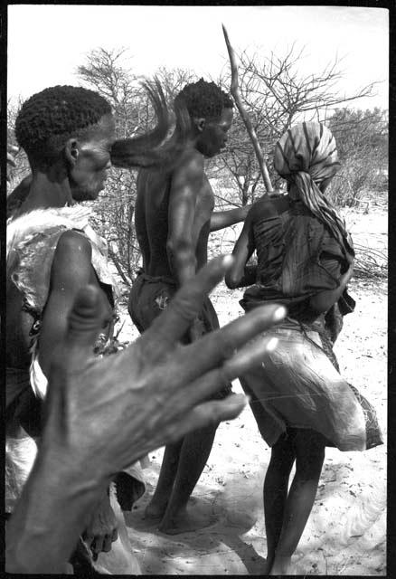 Nǂobe, Tama, and N/kxabe, seen from behind; someone's hand partly obscuring