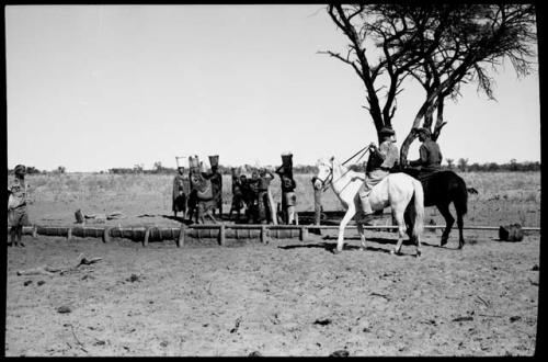 Two men on horses; a group of women by the water trough carrying buckets on top of their heads