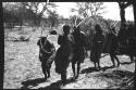 Group of people performing the Eland Dance; !Ani leading a line of men who are holding wooden horns up to their heads
