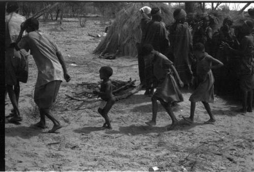 Group of people performing the Eland Dance; group of women clapping; children following the men holding wooden horns to their heads