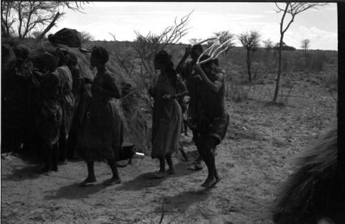 Group of people performing the Eland Dance; group of women clapping; men holding wooden horns to their heads