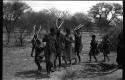 Group of people performing the Eland Dance; !Ani leading a line of people, men holding wooden horns to their heads