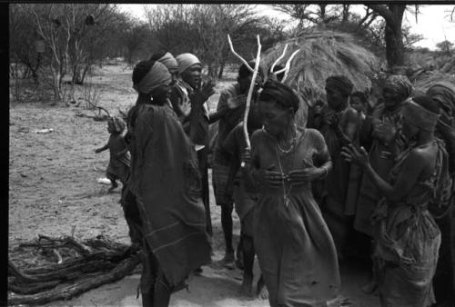 Group of people performing the Eland Dance; group of women clapping as a line of men holding wooden horns to their heads passes by