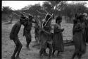 Group of people performing the Eland Dance; group of women clapping; men with wooden horns on their heads