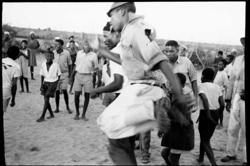 Boy jumping in the air, playing a game in a school yard; people standing and watching in the background