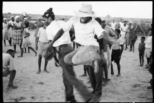 Boy jumping over another boy's leg, playing a game in a school yard; people standing and watching in the background