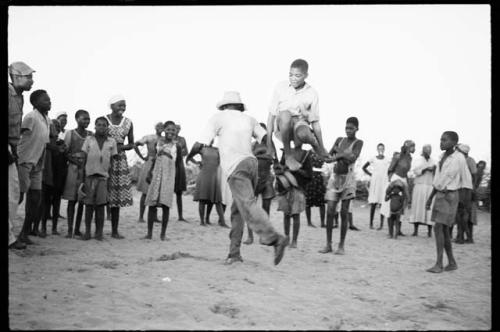 Boy jumping over another boy's leg, playing a game in a school yard; people standing and watching in the background