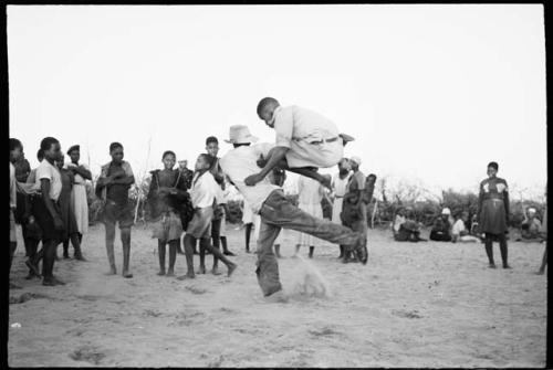 Boy jumping over another boy's leg, playing a game in a school yard; people standing and watching in the background