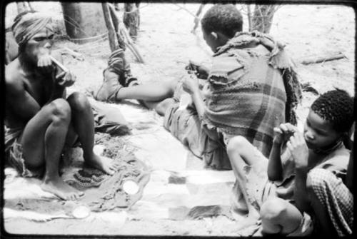 Dadum sitting on animal skins with her husband, Tama, and an unidentified boy. Tama is sewing. (copy of color slide 2001.29.8360)