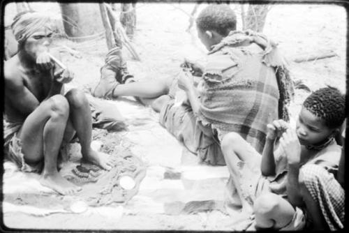 Dadum sitting on animal skins with her husband, Tama, and an unidentified boy. Tama is sewing. (copy of color slide 2001.29.8360)