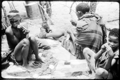 Dadum sitting on animal skins with her husband, Tama, and an unidentified boy. Tama is sewing. (copy of color slide 2001.29.8359)