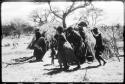 Women performing the Eland Dance, with some standing in front of a skerm, singing and clapping, and others dancing around the skerm (copy of color slide 2001.29.8651)