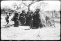 Women performing the Eland Dance, with some standing in front of a skerm, singing and clapping, and others dancing around the skerm (copy of color slide 2001.29.8650)