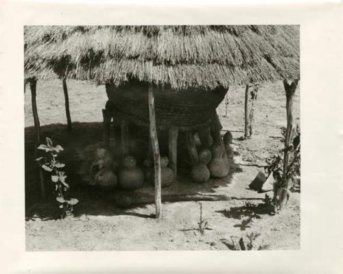 Storage basket supported by wooden struts surrounded by pots, all under a thatched roof (print is a cropped image)