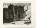 Storage basket supported by wooden struts surrounded by pots, all under a thatched roof (print is a cropped image)