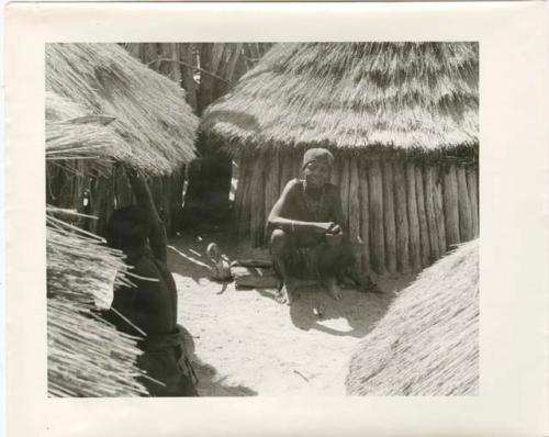 Woman sitting in front of one of several huts (print is a cropped image)