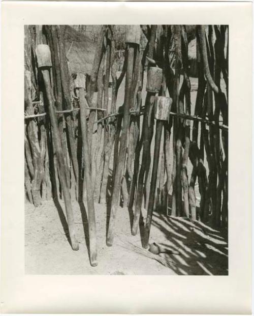 Pestles for pounding grain, leaning against a fence (print is a cropped image)