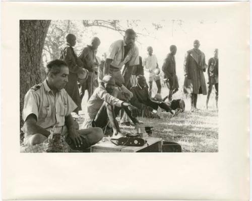 Group of men standing and sitting under a tree, with Senhor Simões in the foreground with a camera (print is a cropped image)