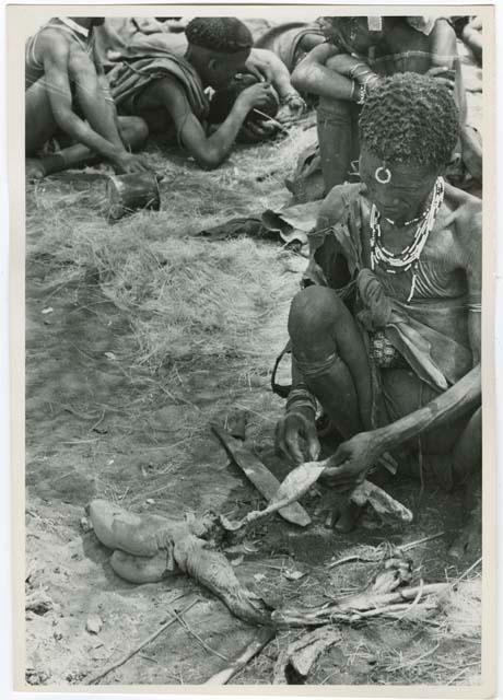 "Old Xama" preparing a piece of skin, with unidentified people in the background  (print is a cropped image)