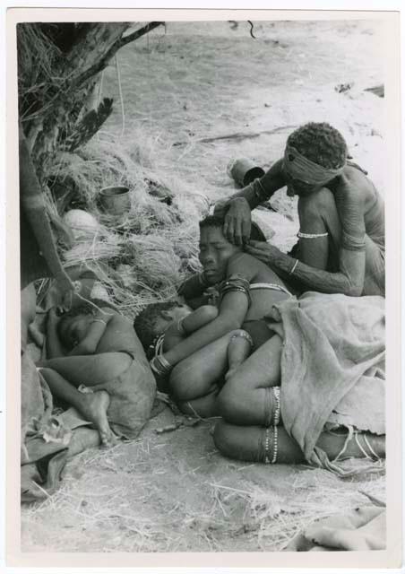 Elderly woman adjusting the earring of a younger woman who is lying with her baby; another child asleep beside them (print is a cropped image)