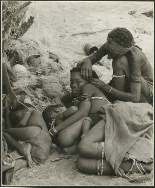 Elderly woman adjusting the earring of a younger woman who is lying with her baby; another child asleep beside them (print is a cropped image)