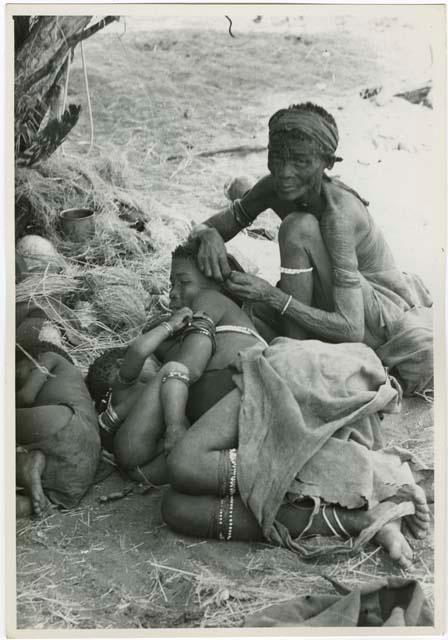 Elderly woman adjusting the earring of a younger woman who is lying with her baby; another child asleep beside them  (print is a cropped image)