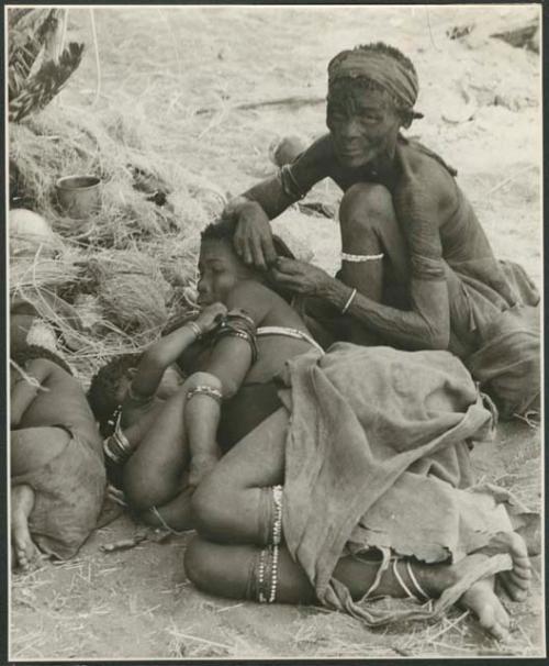 Elderly woman adjusting the earring of a younger woman who is lying with her baby; another child asleep beside them (print is a cropped image)