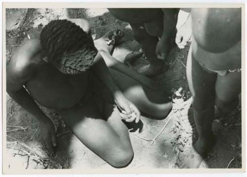 Boy sitting on the ground, with two other people standing beside him (print is a cropped image)