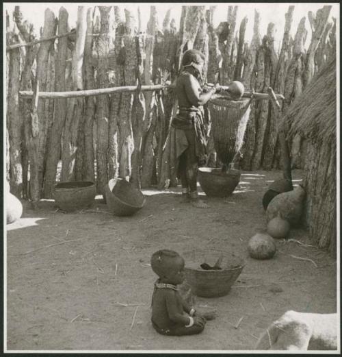 Woman pouring meal through a sieve, and a child sitting on the ground