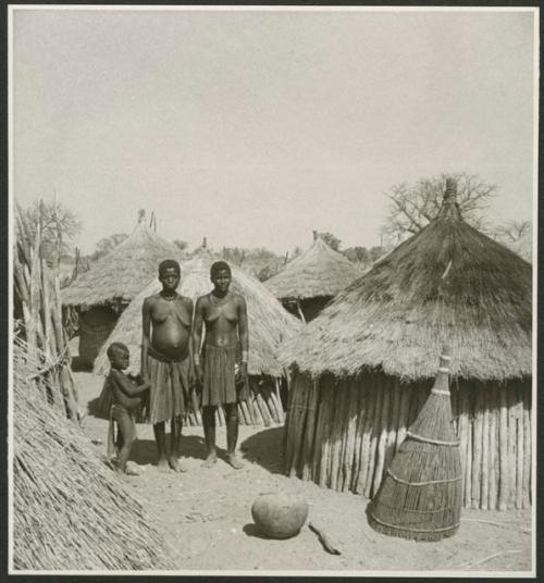 Two women and a child standing next to their huts