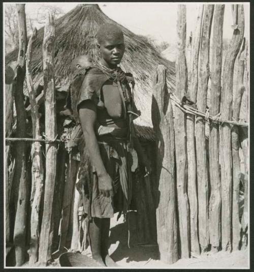 Woman carrying a baby on her back, standing in front of a fence with poles tied with creepers