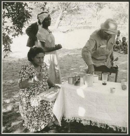 People standing and sitting at camp, with Mm. Simões seated at a table