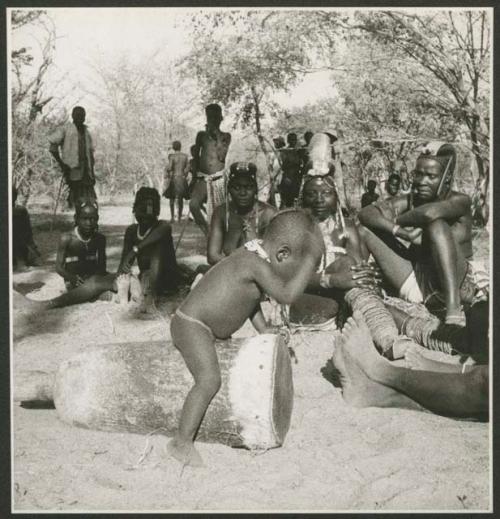 Child playing a drum, and other people sitting and watching