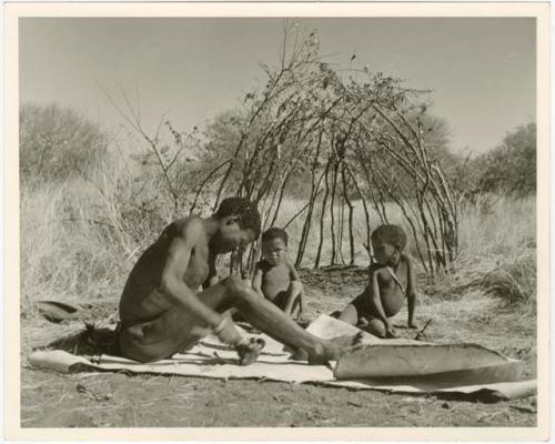 /Ti!kay scraping a skin, and two children sitting and watching, with the frame of a partly-built skerm behind them (print is a cropped image)