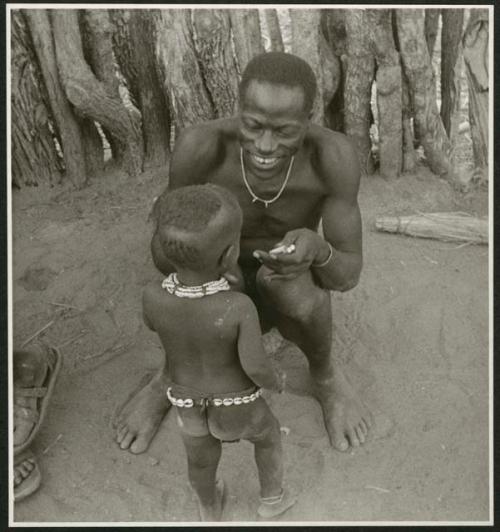 Man sitting, holding cigarettes and showing his filed teeth, and a child standing in front of him