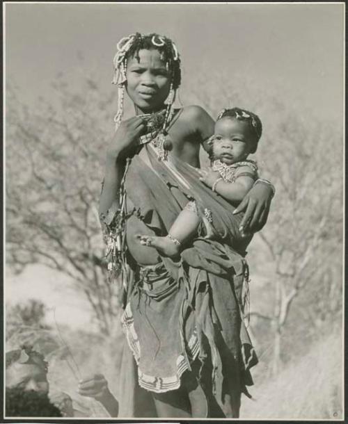 Woman wearing apron and bead hair ornaments, standing and carrying a baby in her kaross (print is a cropped image)