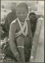 /Gasa (/Qui's wife) wearing an ostrich eggshell necklace; /Qui and another unidentified Ju/'hoan man in the background (print is a cropped image)