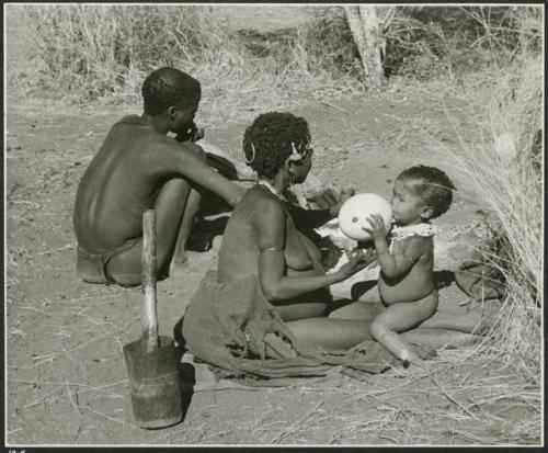 !Ungka (≠Toma's sister) with her daughter, Sa≠gai, sitting on her knees drinking from an ostrich eggshell; Tsamgao (Bau's husband) sitting beside her (print is a cropped image)