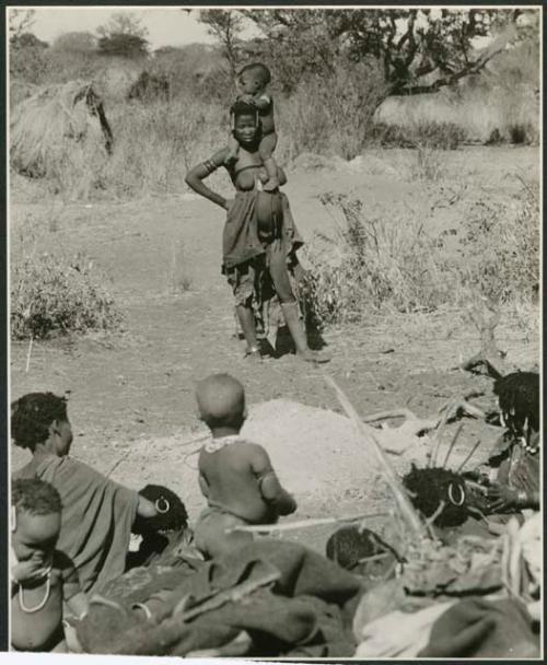 Group of Ju/'hoansi seated around a fire in the foreground; //Kushay ("Gao Helmet's" second wife) standing with her child on her shoulders in the background (print is a cropped image)