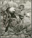 /Naoka, wife of Ti!kay, drinking from an ostrich eggshell, with !Nani behind her, and /Khoa, her daughter, standing beside her (print is a cropped image)