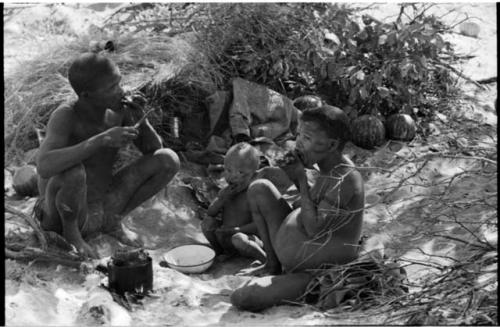 !Gai, Tsekue, and N!whakwe sitting by their fire and eating meat, with meat cooking in a tin on it