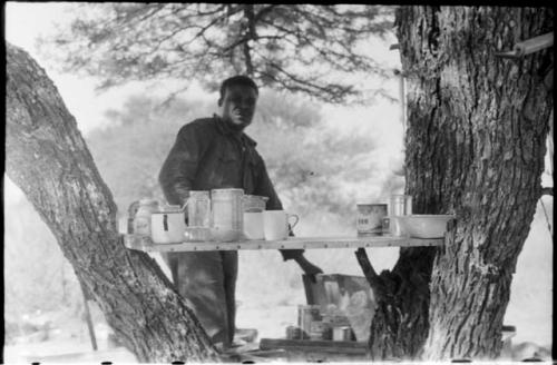 Philip Hameva, expedition cook, standing in his kitchen - a shelf between two tree trunks