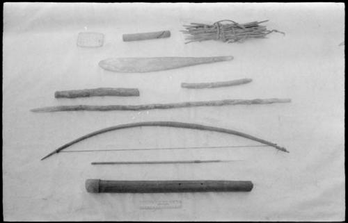 /Twikwe's possessions, including (top to bottom, left to right) a piece of wood hollowed in center used to trip iron traps, a small stick, a bundle of sharpened sticks used to peg down a hide to stretch it, a fire paddle, 2 sticks, a digging stick, a bow, arrow with a bone point shaped like a needle, and a quiver, with a ruler for measurement