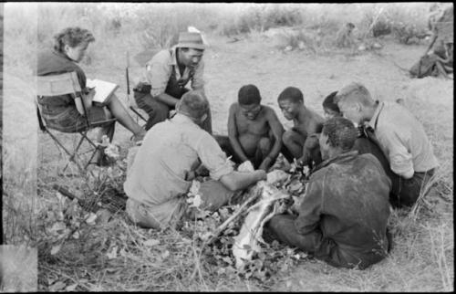 Oukwane and other people sitting and talking to William Donnellan and Elizabeth Marshall Thomas, with Wilhelm Camm interpreting. Oukwane is naming the internal parts of an animal.