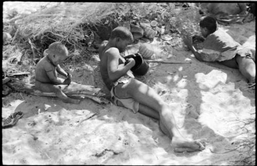 !Gai lying down in his living place, and N!whakwe sitting on a log and playing with a knife next to him