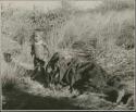 Woman lying on the ground wrapped in kaross, with Khuan//a, daughter of //Ao and ≠Nisa, standing next to her (print is a cropped image)