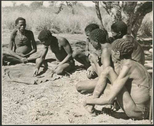 Group of men sitting under a big tree, throwing and watching oracle disks; the thrower pointing at one disk and reading it (print is a cropped image)