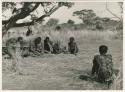 Group of men sitting under a tree, throwing and reading oracle disks, from a distance (print is a cropped image)